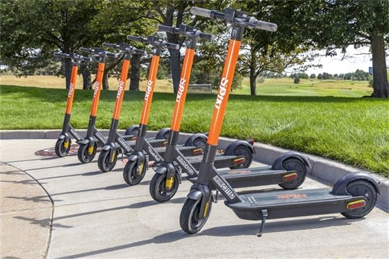 Six spin electric scooters parked in on sidewalk near a park